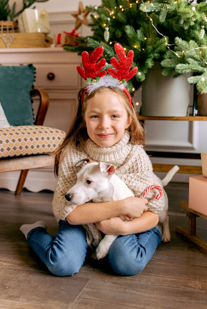Smiling girl with reindeer headband hugging dog by Christmas tree indoors.